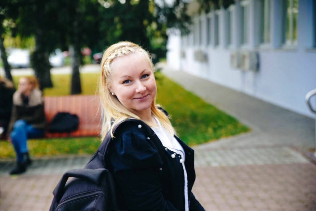 The picture shows Ljiljana with a backpack. She smiles at the camera and in the background there is a park bench and a piece of meadow.